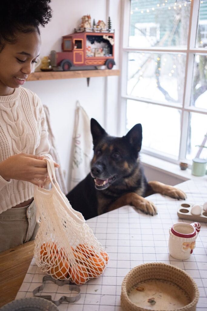 A woman with a dog in a cozy kitchen setting, holding a bag of oranges.